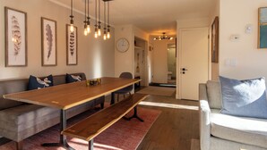 En moderne stue med et træspisebord, bænke, en grå sofa og en grå lænestol. Vægophængte fjer-kunstværker, et ur og en hængende lampe.