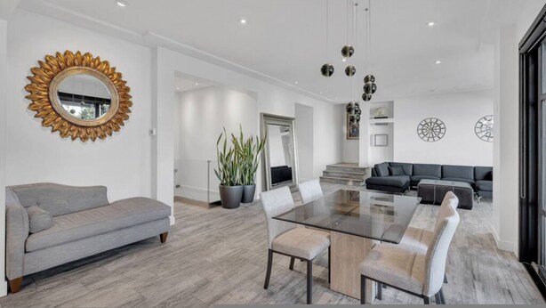 A modern living room with a glass dining table, grey sofas, and a large mirror with a sunflower frame. - Sunset Lane Waterfront Home (West Vancouver)