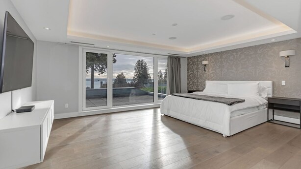 A modern bedroom with a large sliding glass door leading to a balcony, a bed with white bedding, a bedside table, and a TV mounted on the wall. - Ocean City View Home (West Vancouver)