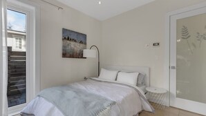 A bedroom with a bed, a nightstand, a lamp, a painting, a sliding glass door, and a small round table. - Ocean City View Home (West Vancouver)