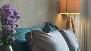 En pænt redt seng med turkis sengetøj, en vase med lilla blomster, en lampe og en bog på et natbord.