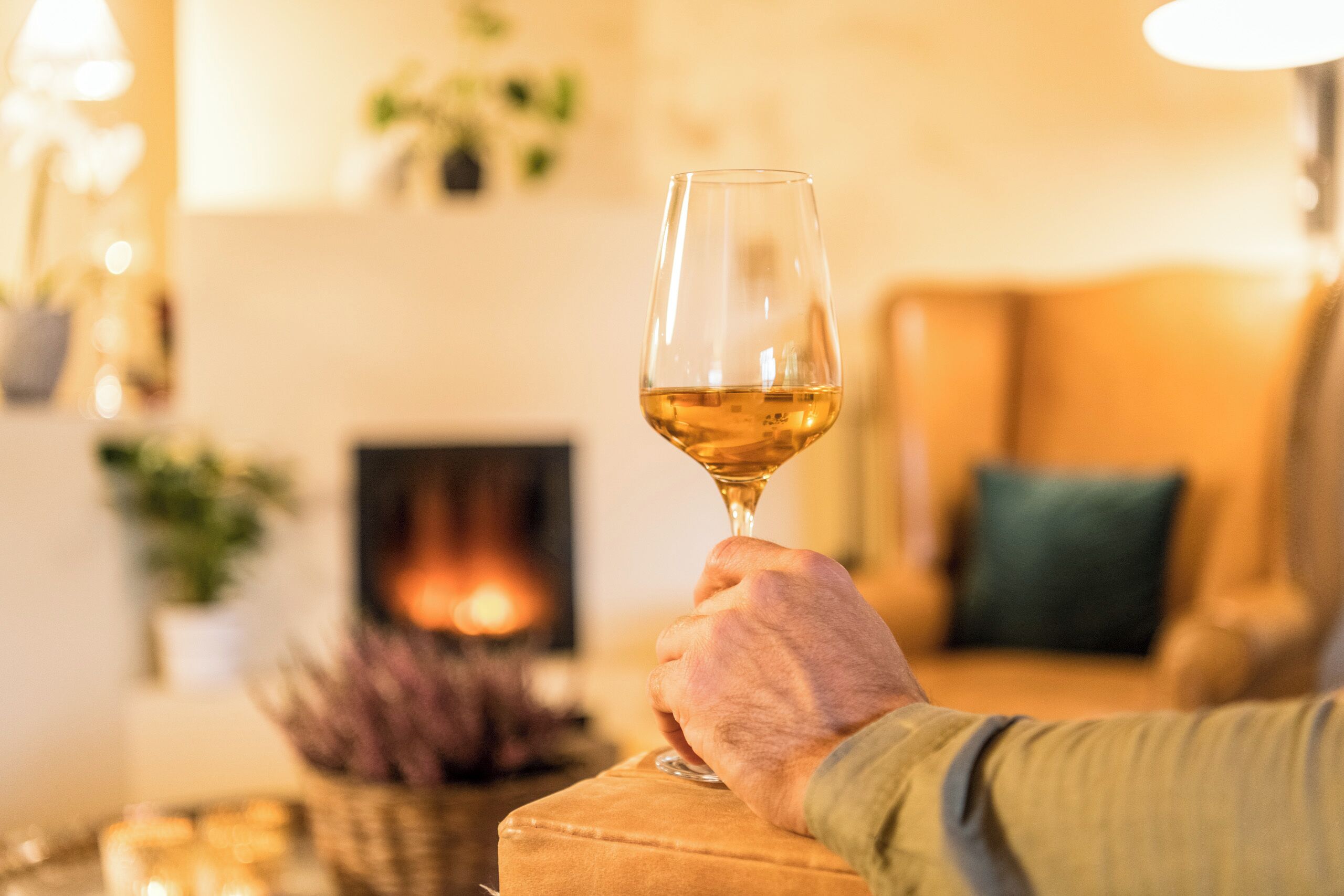 Une main tenant un verre de vin dans une pièce chaleureusement éclairée, avec une cheminée en arrière-plan.