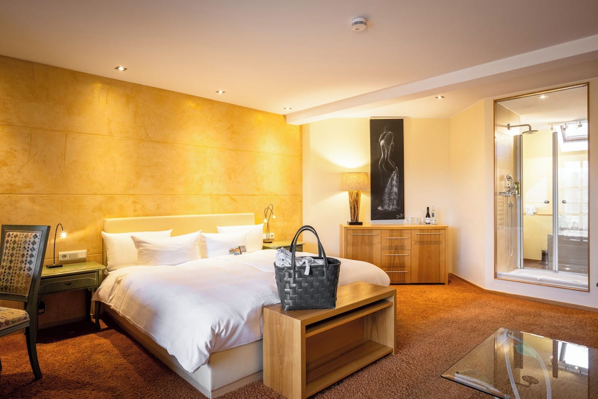 Habitació d'hotel moderna amb un llit gran, una tauleta de nit de fusta, un escriptori, una taula de cafè de vidre, un bany amb dutxa de vidre i un mirall gran.