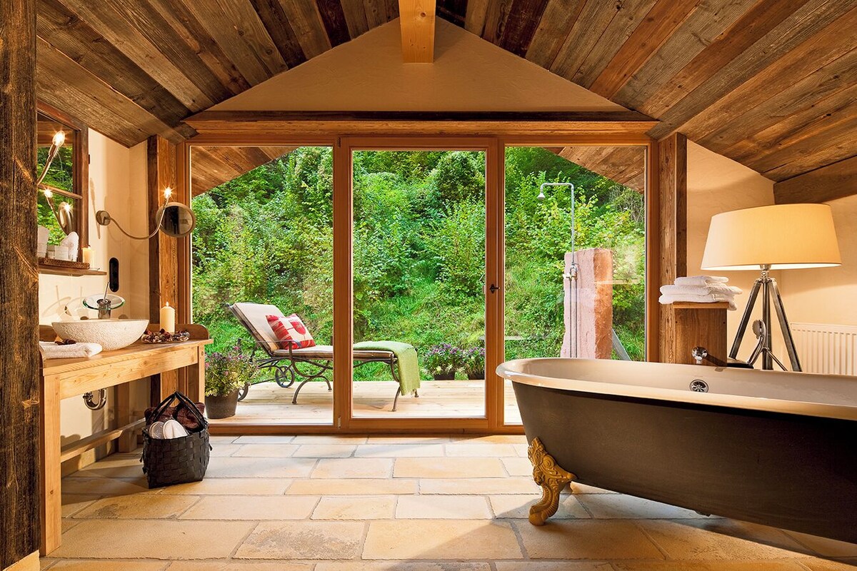 Un baño con una bañera grande, un lavamanos y una silla cerca de un ventanal corredizo que da a un jardín.