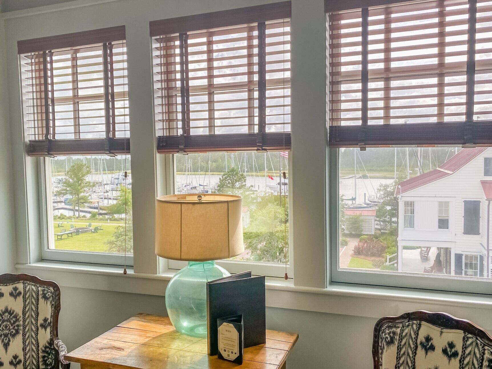 A room with three windows, each with wooden blinds, a table with a lamp and a framed certificate, and a view of a marina and a house across the water.