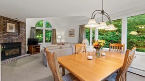 A cozy living room with a fireplace, a television, and a sofa. There is a dining area with a wooden table and chairs, and large windows offering a view of greenery outside. - Fall Line Condo D6 3 Bedroom Condo (Killington)