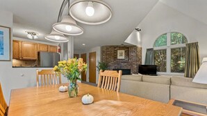 A dining area with a wooden table, chairs, and a vase of flowers. There is a kitchen in the background with wooden cabinets and a refrigerator. A living area with a sofa and a television is visible through a window. - Fall Line Condo D6 3 Bedroom Condo (Killington)