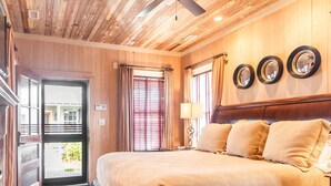A hotel room with a wooden ceiling, a large bed with beige bedding, a nightstand with a lamp, and a door leading outside.