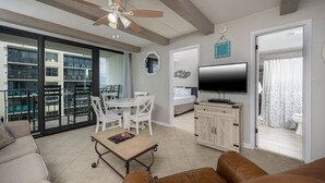 A spacious living room with a sofa, a dining table with chairs, a television, and a balcony with a view of the city. - Island Winds East 610 2 Bedroom Condo (Gulf Shores)
