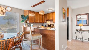 A kitchen with wooden cabinets, a dining area with wicker chairs, and a view of the outdoors through a window. - Maui Kamaole, #a-106 1 Bedroom Condo (Kihei)