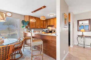 A kitchen with wooden cabinets, a dining area with wicker chairs, and a view of the outdoors through a window. - Maui Kamaole, #a-106 1 Bedroom Condo (Kihei)