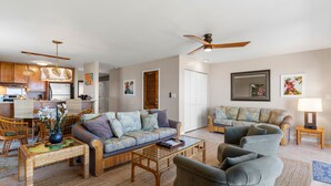 A spacious living area with a sofa, armchairs, and a coffee table. There is a kitchen area with cabinets and a refrigerator. The room has a ceiling fan and wall-mounted artwork. - Maui Kamaole, #a-106 1 Bedroom Condo (Kihei)