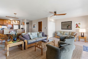 A spacious living area with a sofa, armchairs, and a coffee table. There is a kitchen area with cabinets and a refrigerator. The room has a ceiling fan and wall-mounted artwork. - Maui Kamaole, #a-106 1 Bedroom Condo (Kihei)