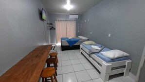Ein kleiner, kompakter Raum mit einem Holztisch und vier Stühlen, einem Einzelbett mit blauer Decke sowie einer weißen Klimaanlage an der Wand.