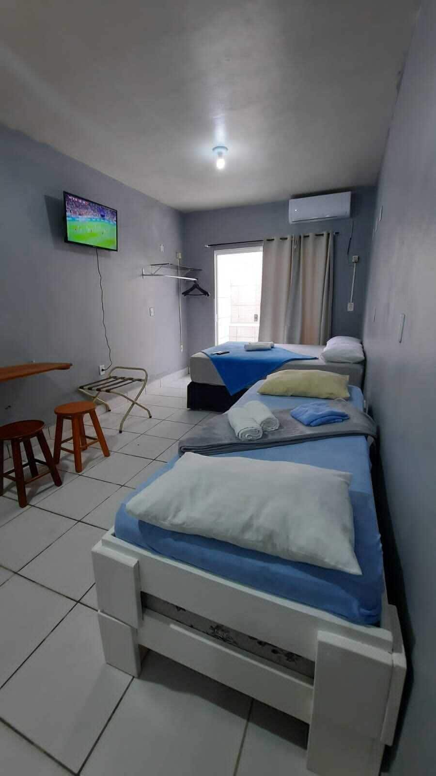 A hotel room with a bed, a TV, a window with curtains, and a small stool.