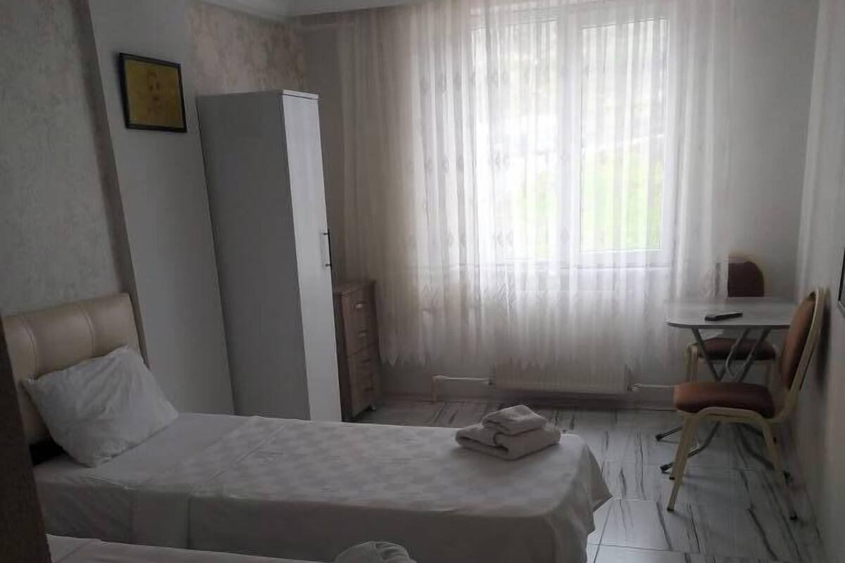 A hotel room with two beds, a desk, a chair, a wardrobe, and a window with sheer curtains.
