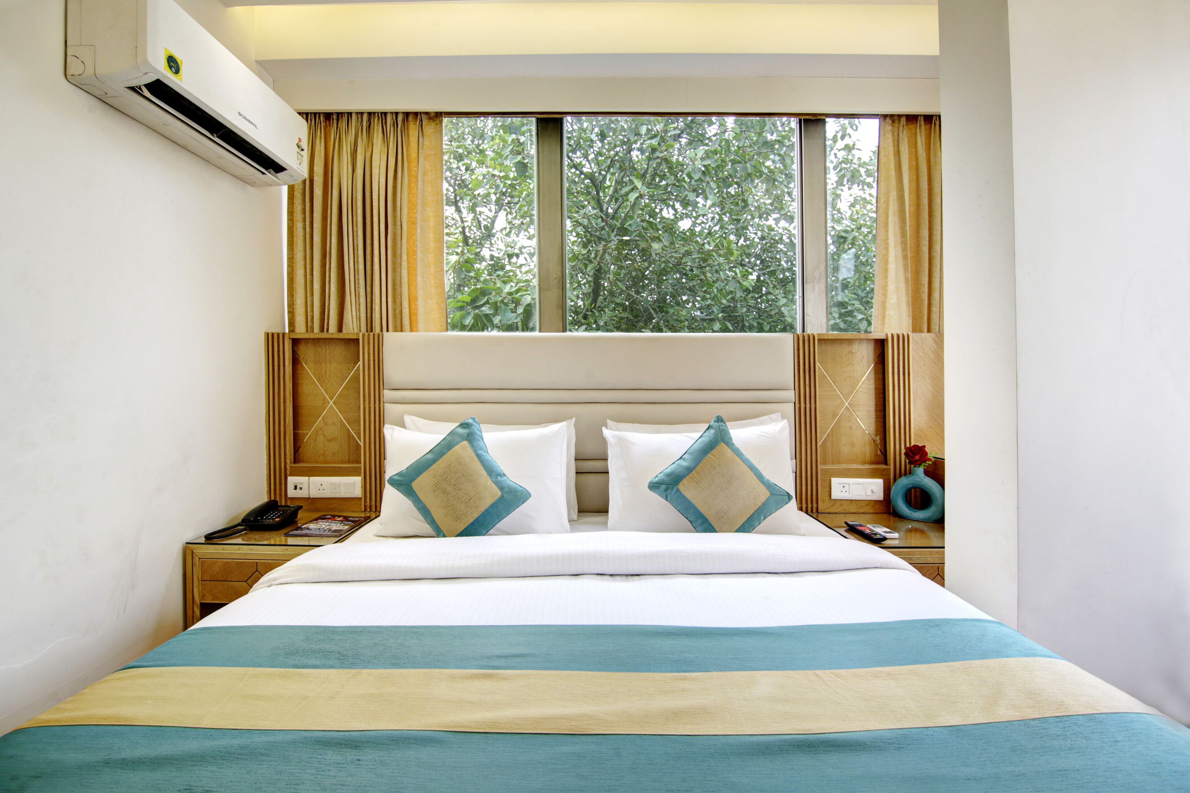 Habitación de hotel con una cama grande, dos mesitas de noche, aire acondicionado, ventana con cortinas y vista a árboles.