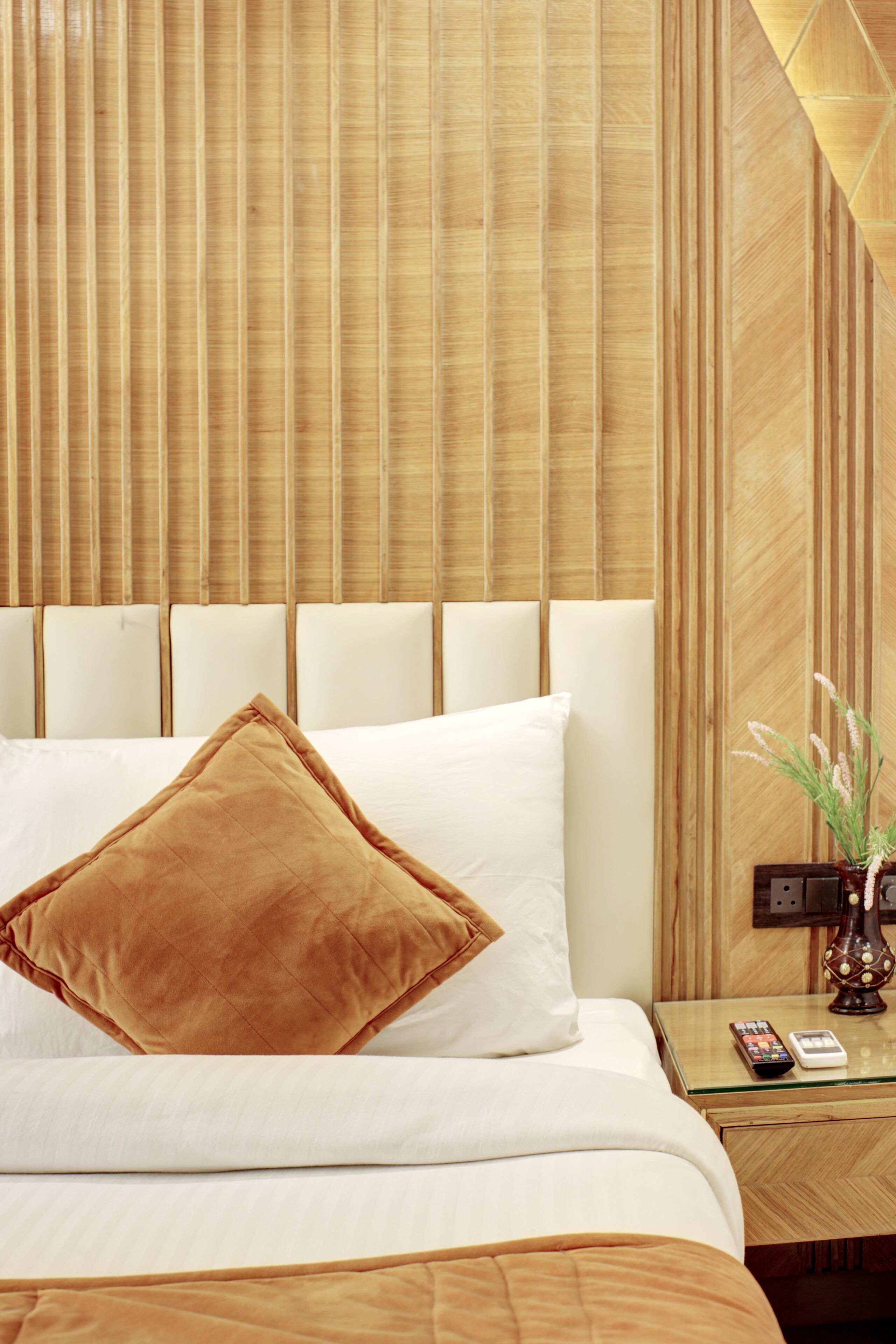A neatly made bed with white and brown bedding, a wooden headboard, and a bedside table with a remote control and a vase with flowers.