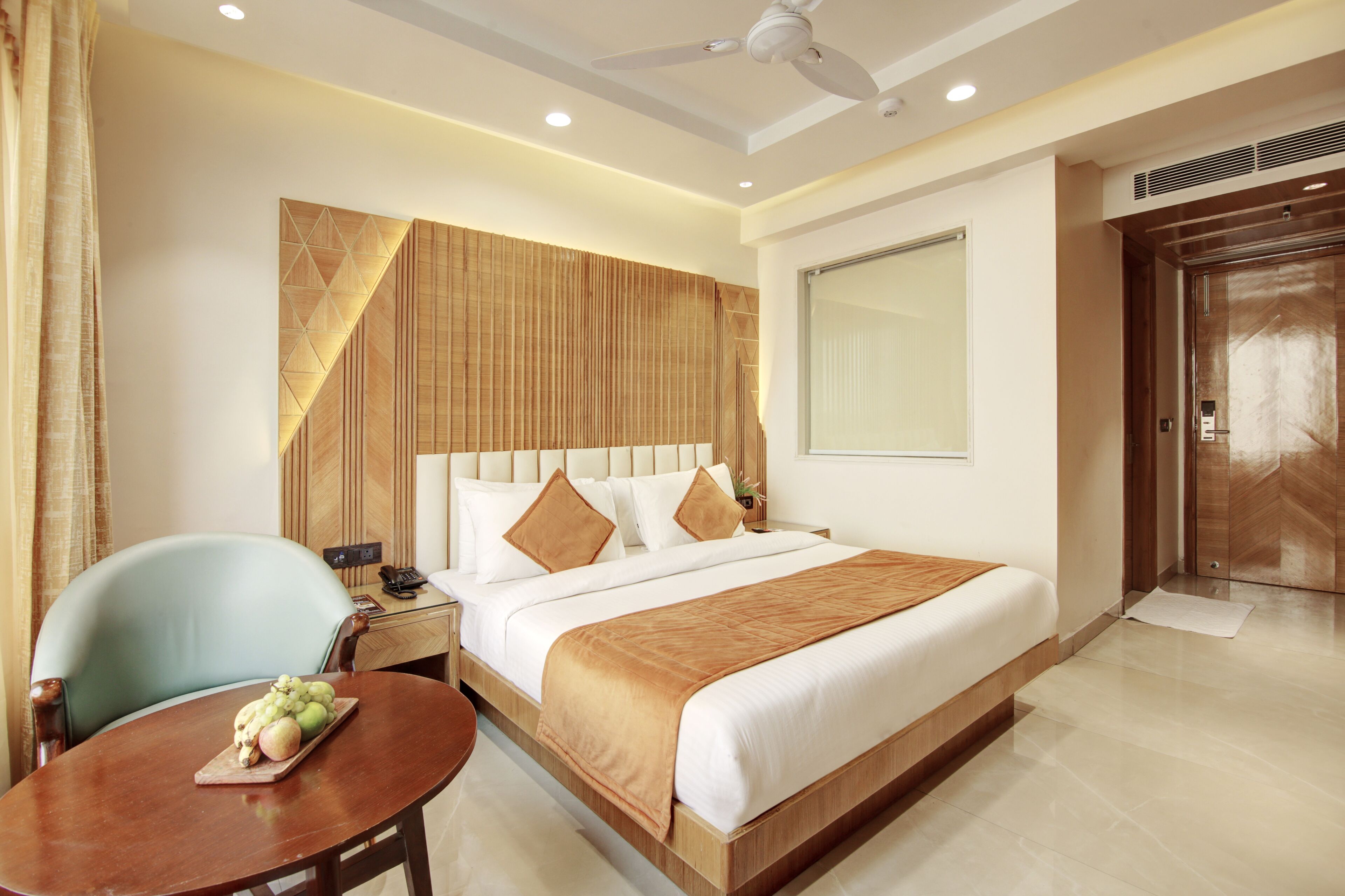 A hotel room with a large bed, a wooden headboard, a desk with a chair, and a dining table with fruit.