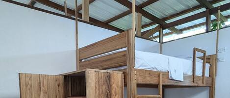 A bunk bed with a wooden ladder and a shelf, under a corrugated metal roof, in a room with tiled flooring.