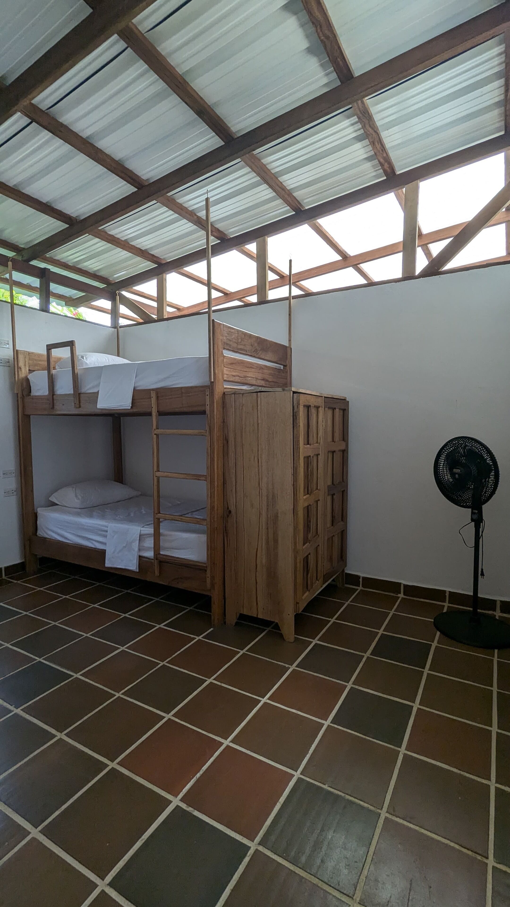 Una habitación con una cama litera, un armario de madera y un ventilador de pie.