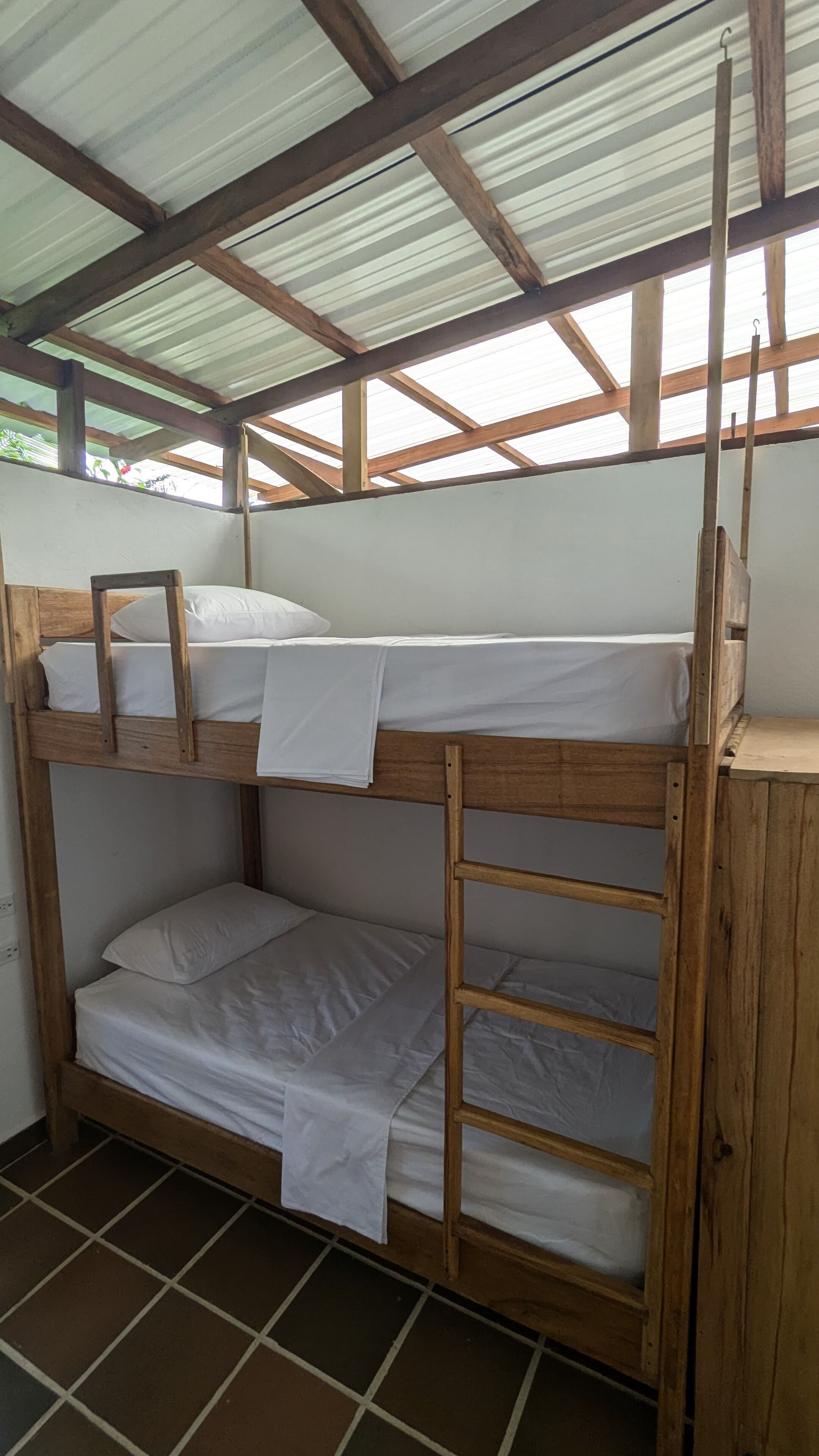 A bunk bed with a wooden frame and white bedding, under a corrugated metal roof.