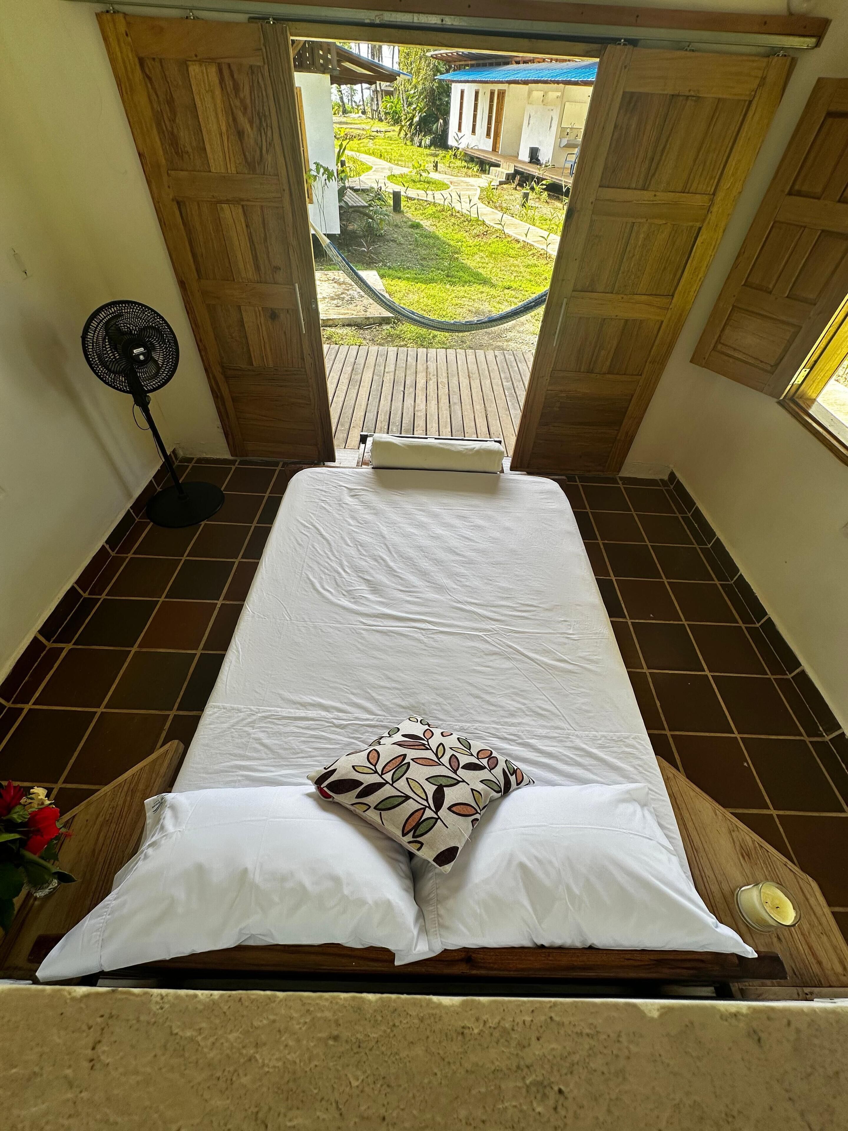 Quarto com cama, ventilador e vista para um jardim através de portas abertas.