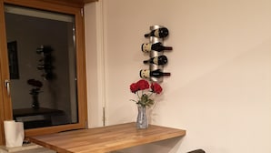 Een klein eetgedeelte met een houten tafel, twee zwarte barkrukken, een vaas met rode bloemen en een wandmontage wijnrek.
