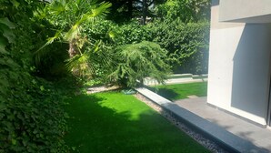Uitzicht op een verzorgde tuin met verschillende bomen en struiken, een strak gemaaid gazon en een geplaveid pad.