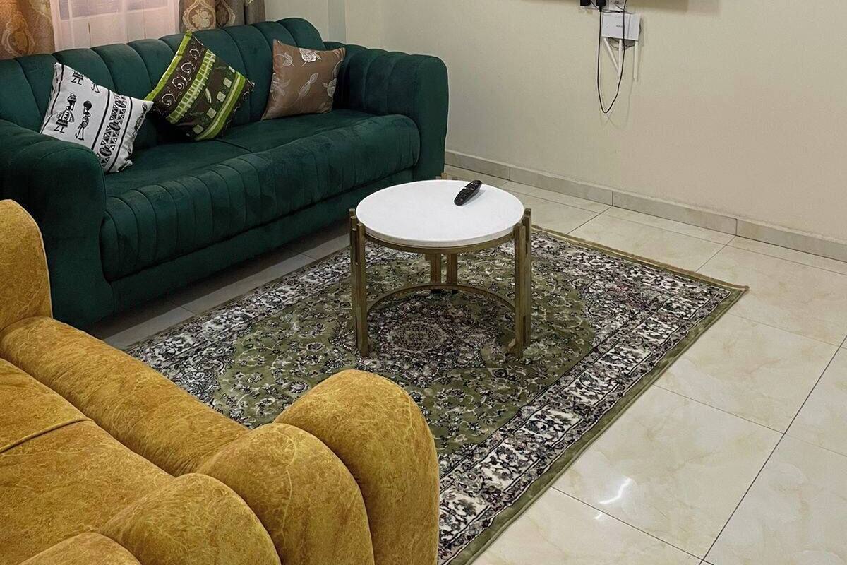 Uma sala de estar com um sofá verde, uma poltrona amarela, uma mesa pequena branca e uma televisão de tela plana na parede.