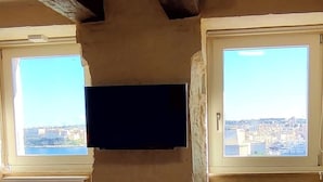 A room with a television mounted on the wall between two windows, each with a view of a cityscape.