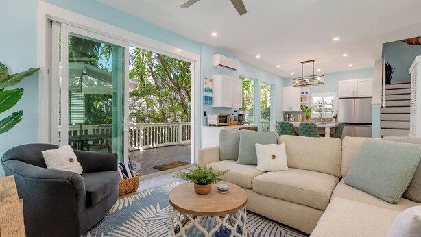A spacious living area with a sofa, armchairs, and a coffee table. There is a sliding glass door leading to a balcony with a view of greenery.