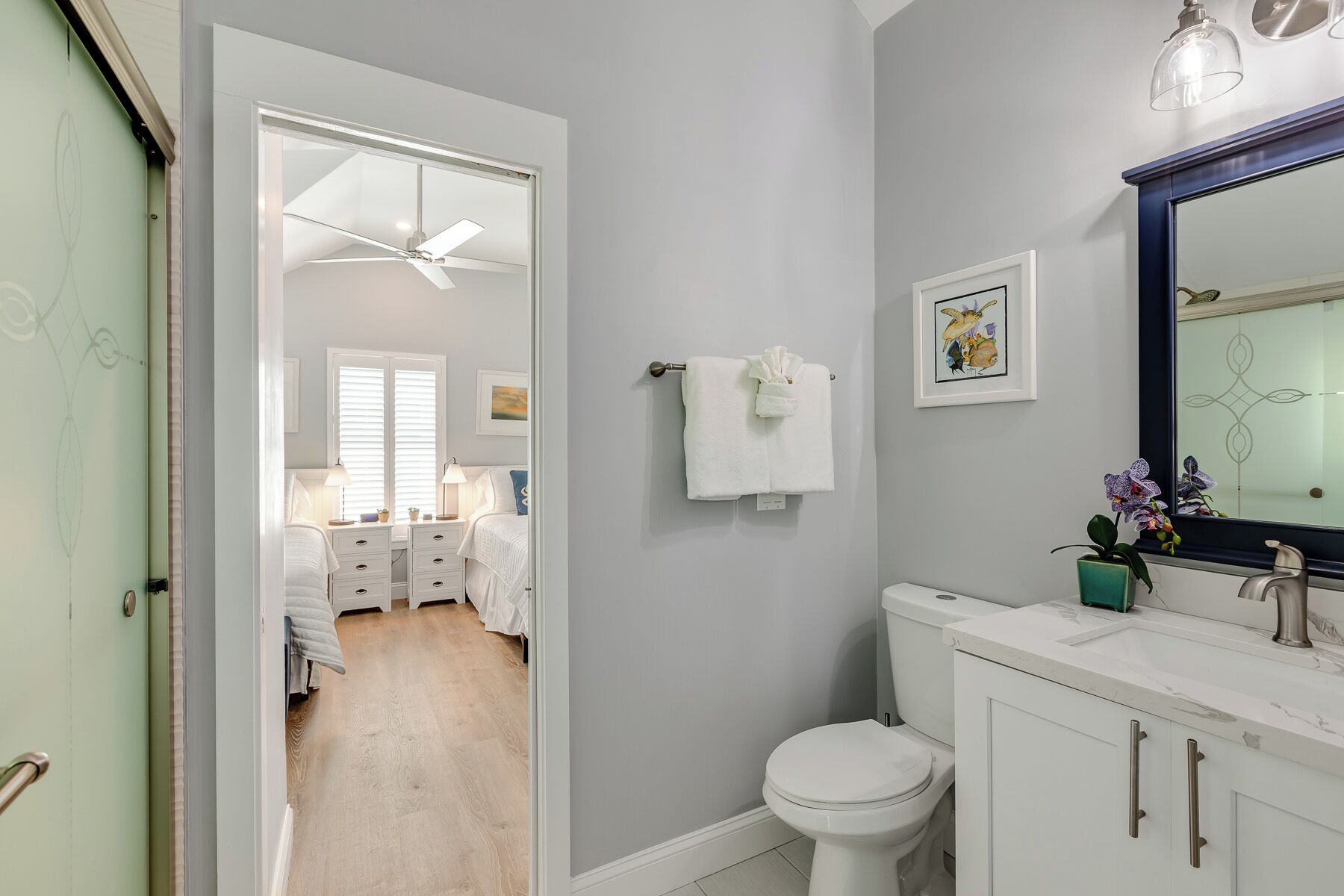 Une salle de bain avec des toilettes, un lavabo et un miroir. Une serviette est accrochée au mur et un ventilateur de plafond est installé au-dessus des toilettes.