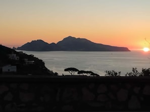 Vista de un atardecer costero con montañas al fondo y un muro de piedra en primer plano.