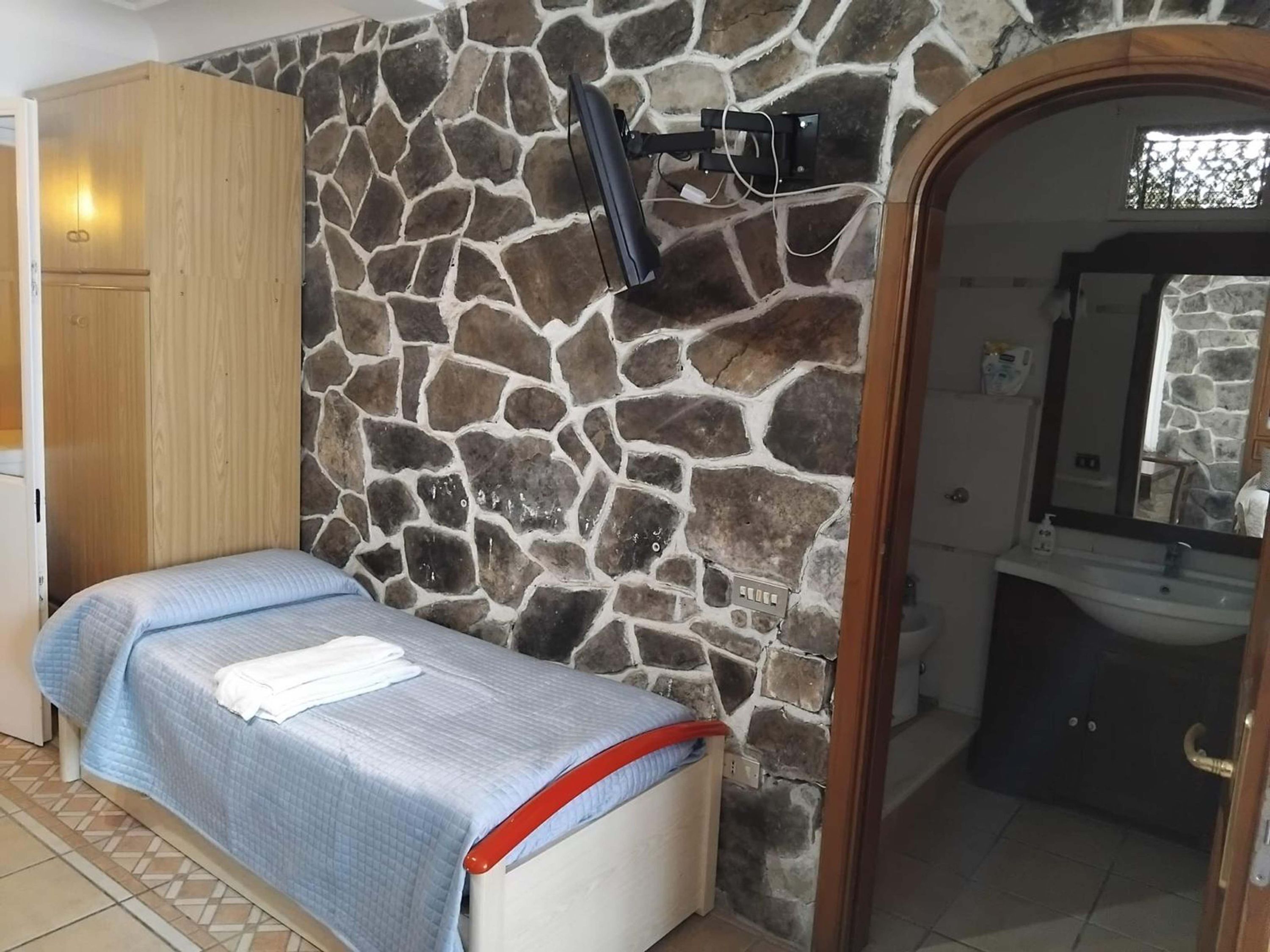 A bedroom with a stone wall, a bed with a blue quilt, a wooden wardrobe, and a bathroom with a sink and mirror.