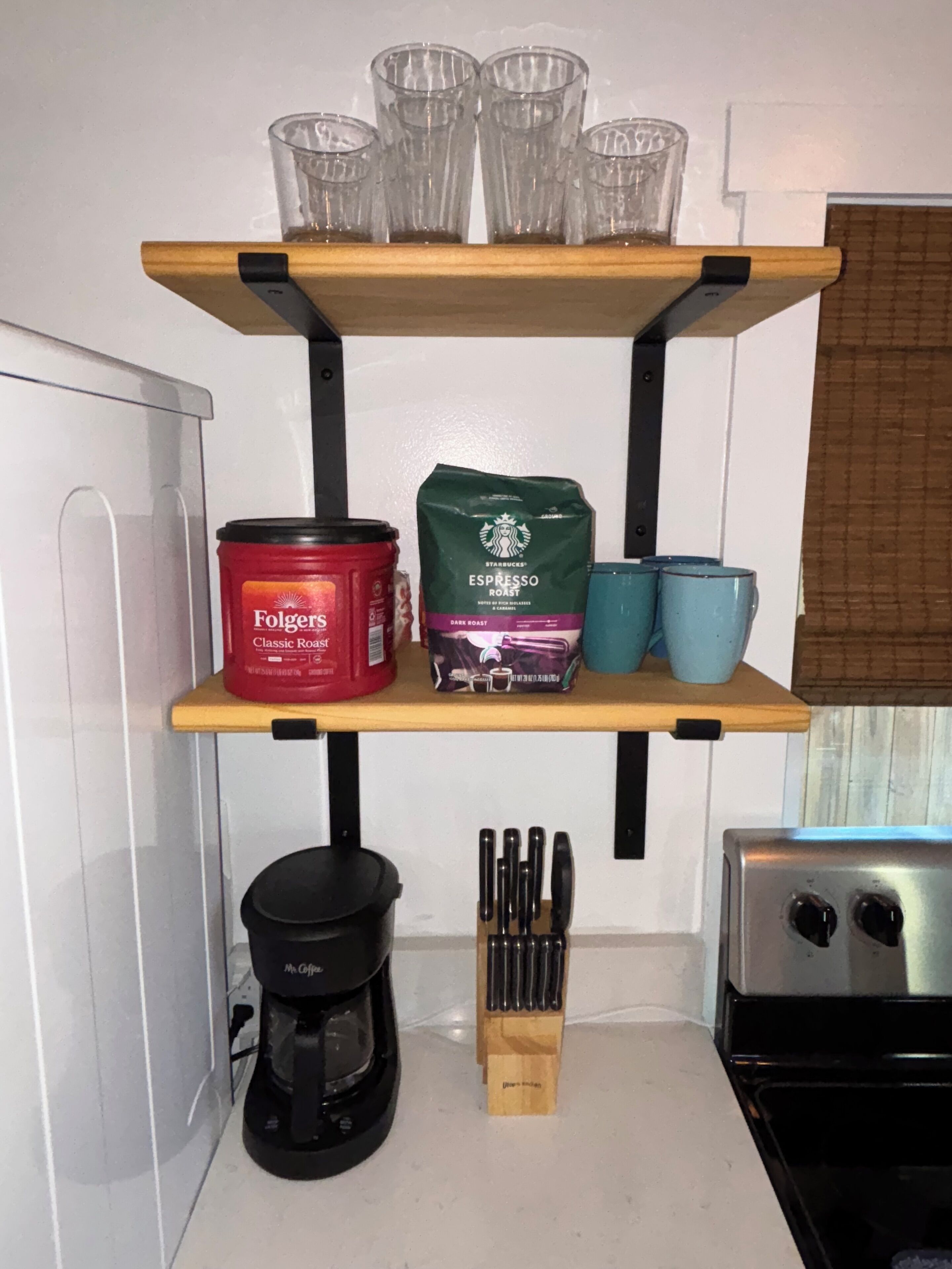 Une étagère de cuisine avec une machine à café, un bocal de café moulu Folgers Classic Roast, un paquet de café moulu Starbucks Espresso Roast et deux tasses bleues.