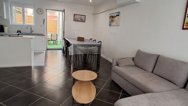 A modern living room with a grey sofa, a dining area with a table and chairs, and a kitchen area with a sink and stove.