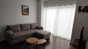 A living room with a grey sectional sofa, a small round coffee table, a TV on a wooden stand, and a window with patterned curtains.