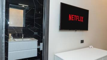 A modern bathroom with a black marble vanity, a white sink, and a wall-mounted television displaying 'NETFLIX'.