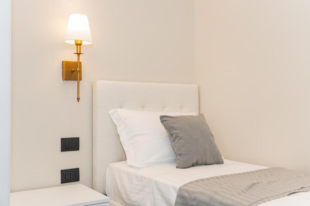 A hotel room with a white bed, a grey pillow, a white nightstand, a wall-mounted lamp, and two switches on the wall.