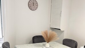 Un coin repas avec une table recouverte d’une nappe à motifs, quatre chaises grises, un vase contenant des plantes séchées et une horloge murale.