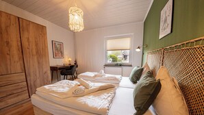 A bedroom with two beds, wooden headboard, a desk with a chair, a window with blinds, and a hanging light fixture.