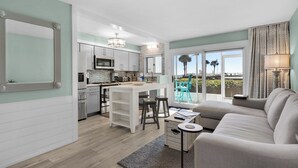 A modern kitchen with white cabinets, stainless steel appliances, and a central island. There is a dining area with a white table and two bar stools. A grey sofa and a small round table are in the living area. - Sand Cliffs 108: Steps to beach & pool access + Free Attraction Tickets! (Seacrest Beach)
