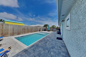 Una piscina rettangolare con bordo piastrellato, recinzione in legno e una casa con parete strutturata.