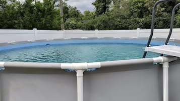 Piscina rectangular elevada con borde de azulejos blancos y azules, rodeada por una valla blanca y con árboles y cielo nublado de fondo.