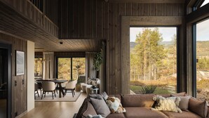 Un intérieur moderne avec un plafond en bois, un canapé d’angle, un coin repas et une grande fenêtre donnant sur la forêt.