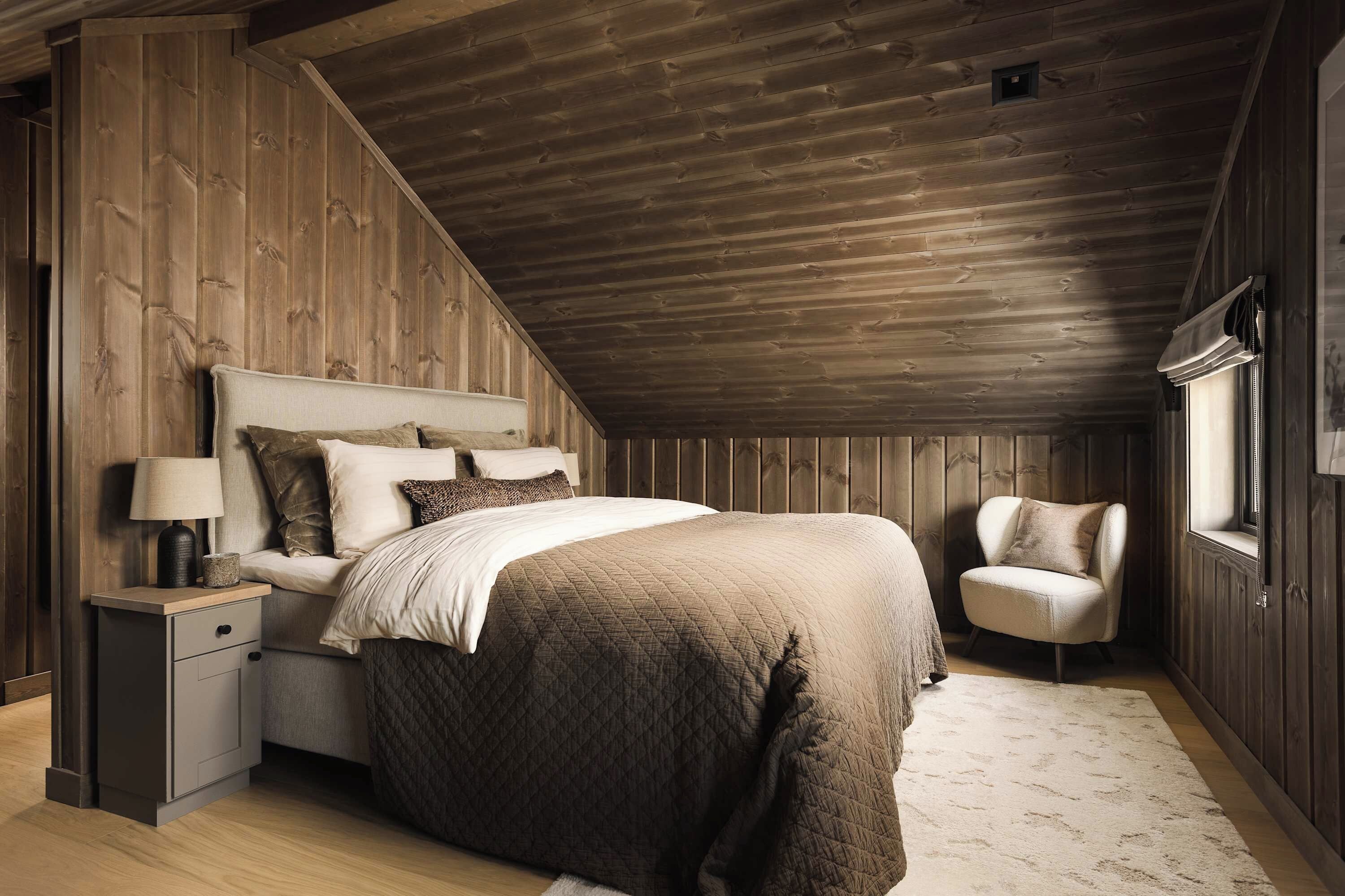 Une chambre avec un plafond en pente, un lit recouvert d’une courtepointe matelassée, une table de chevet avec une lampe et une chaise.