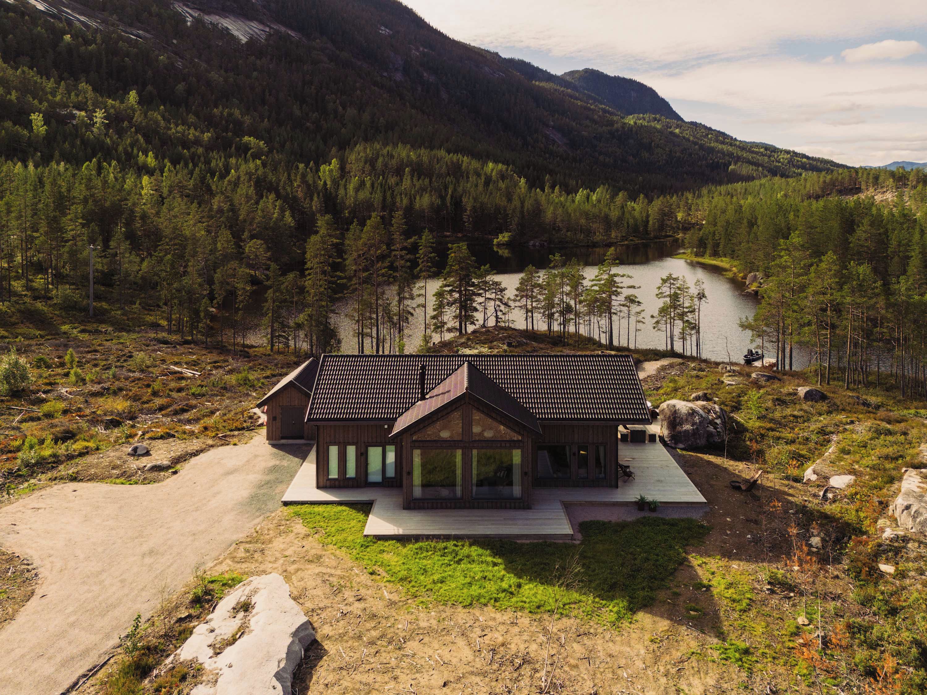 A modern cabin with a large deck, surrounded by a forest and a lake.