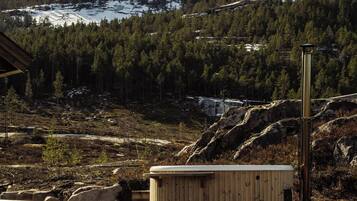 Una terraza de madera con un brasero, una pequeña construcción de madera y vistas a una cadena montañosa nevada.