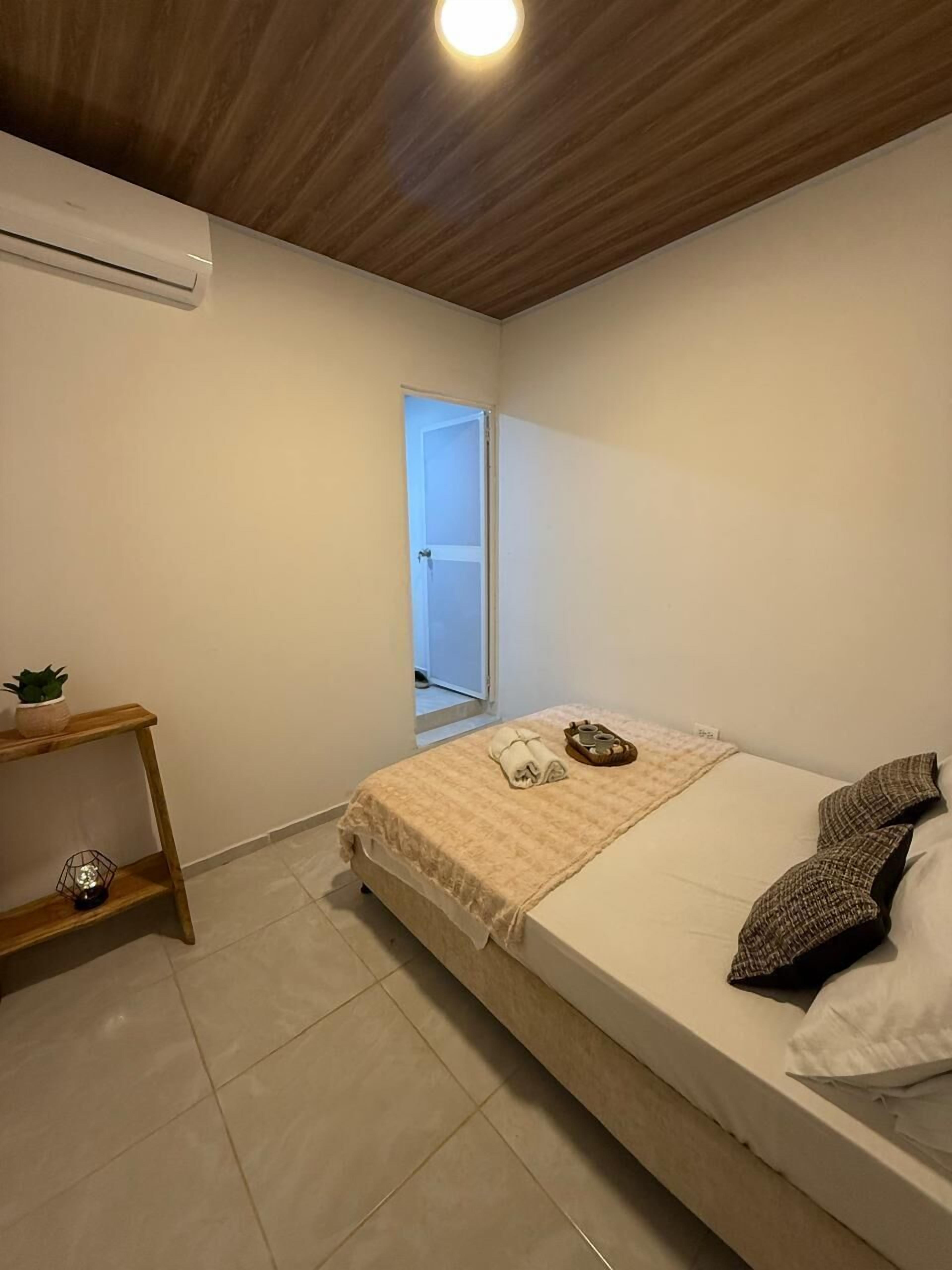 Een slaapkamer met een bed, een nachtkastje met een plant, een raam en een plafondlamp.